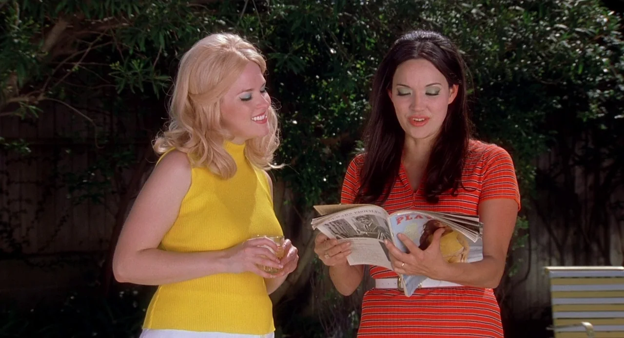 Viva (2007) movie still: medium — Two women stand in front of dark green bushes on a sunny day. The blonde woman holds a glass while the dark-haired…