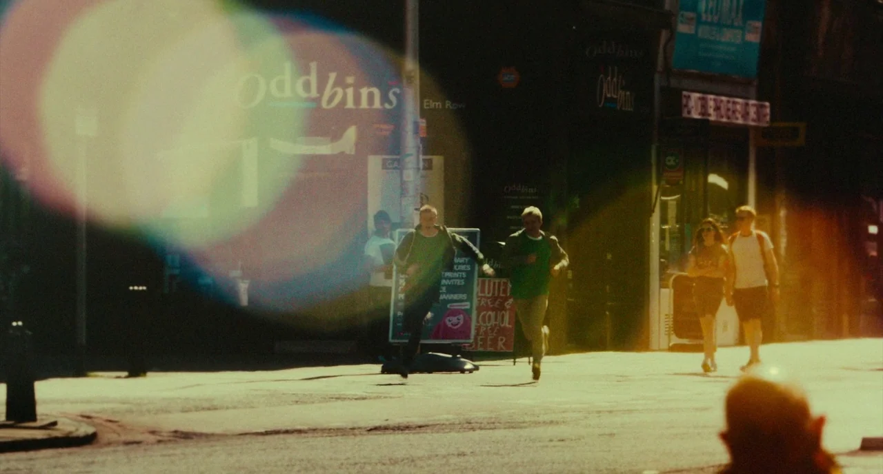 T2 Trainspotting (2017) movie still: wide — An urban street with storefronts featuring signage, illuminated by low-angle golden hour sunlight. Two…