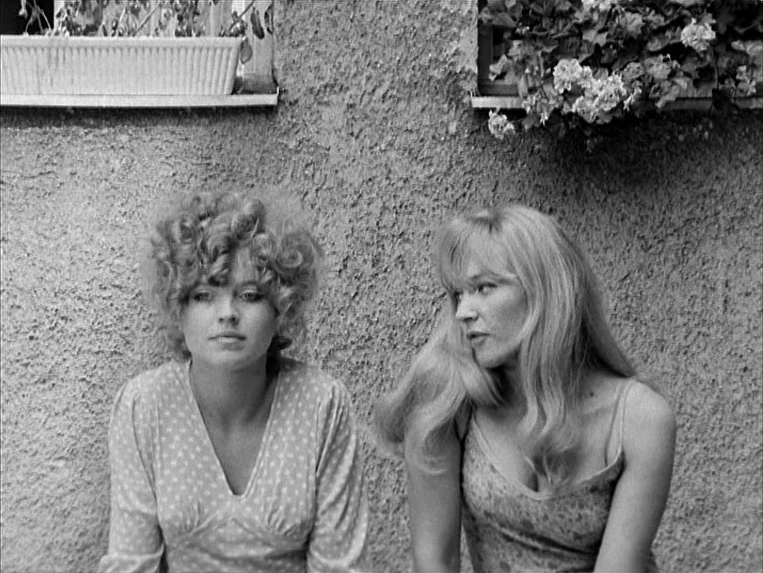 Katzelmacher (1969) movie still: medium — Two women sit positioned against a textured concrete wall with flower boxes above them. The woman on the…
