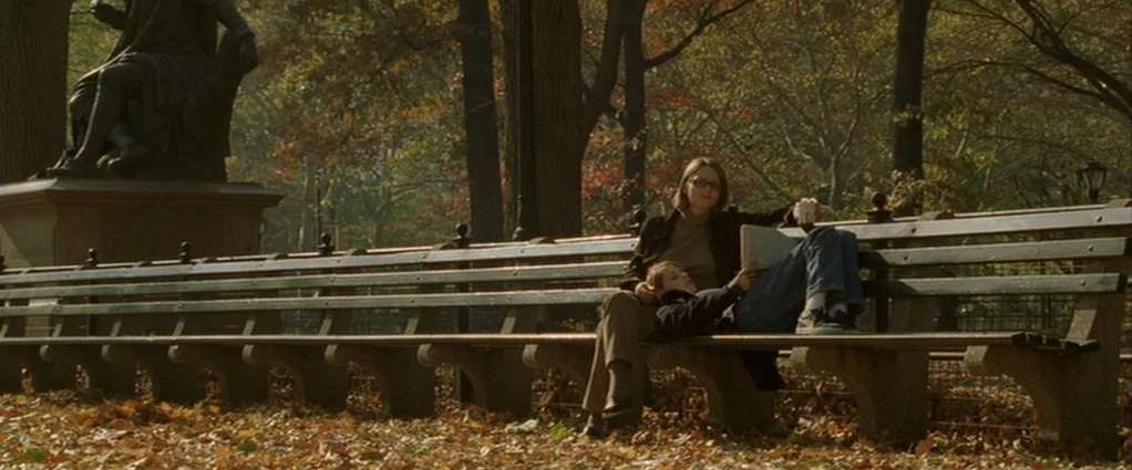 Panic Room (2002) movie still: wide — A park bench sits under autumnal trees covered in fallen leaves next to a statue. A woman reads a book while a…