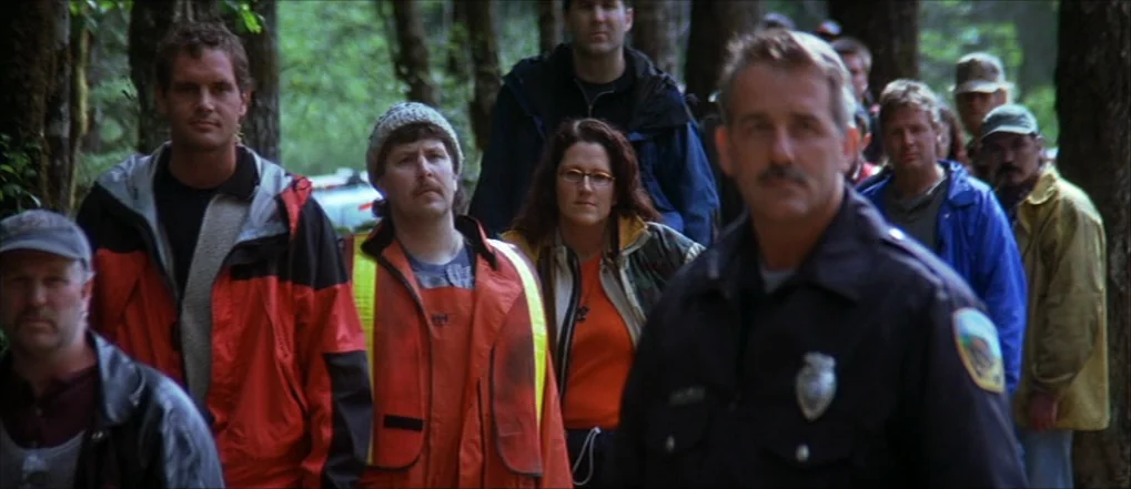 Insomnia (2002) movie still: medium — A diverse group of people stand in a line within a dense wooded area. They wear varied outdoor clothing and…