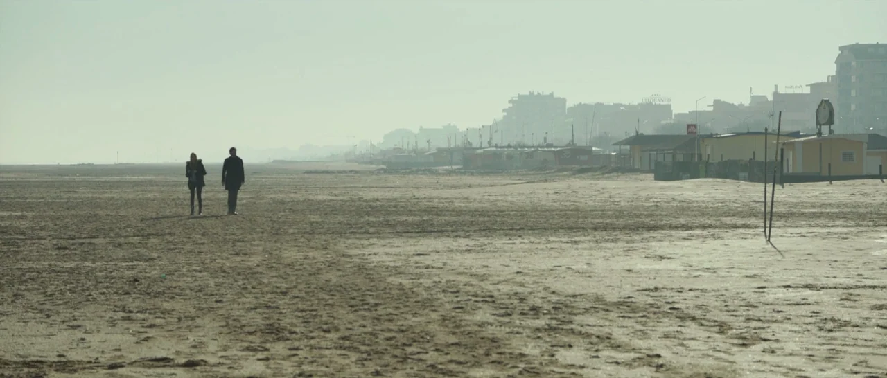 The Face of an Angel (2014) movie still: wide — A vast, flat, overcast beach borders a line of low-rise buildings in the distance. Two figures walk…