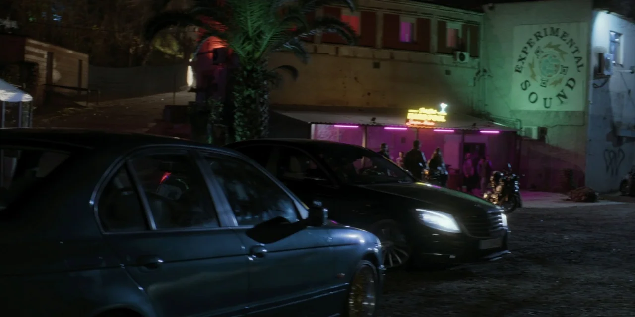 Your Fault (2024) movie still: wide — A night scene shows a gravel parking lot with parked cars in the foreground. A small building with an…