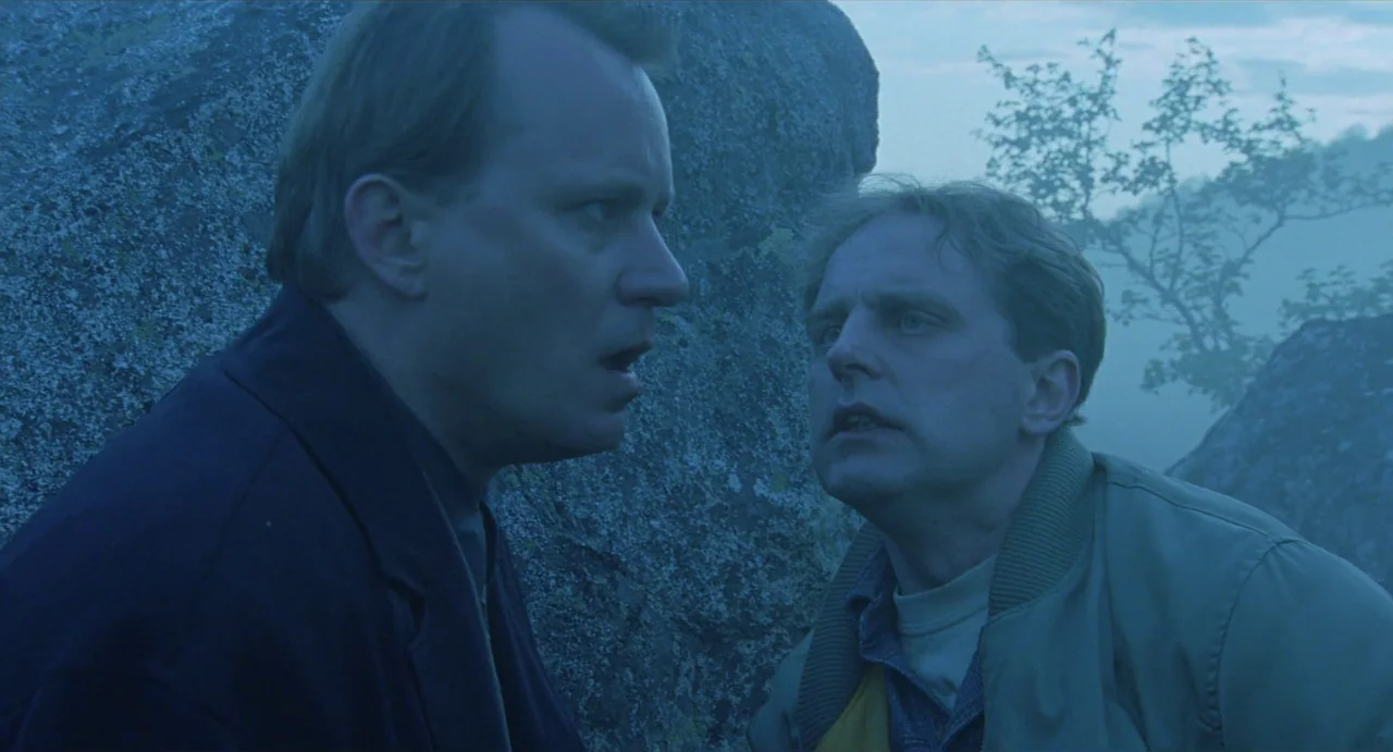 Insomnia (1997) movie still: medium — Two men stand close together against a large gray boulder on a hillside. The background is partially obscured…