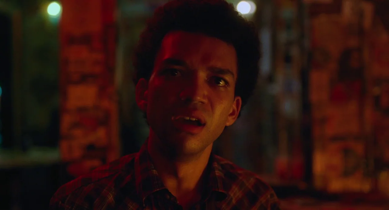 I Saw The TV Glow (2024) movie still: closeup — A dimly lit interior space with dark walls and out-of-focus background textures. A young man with…