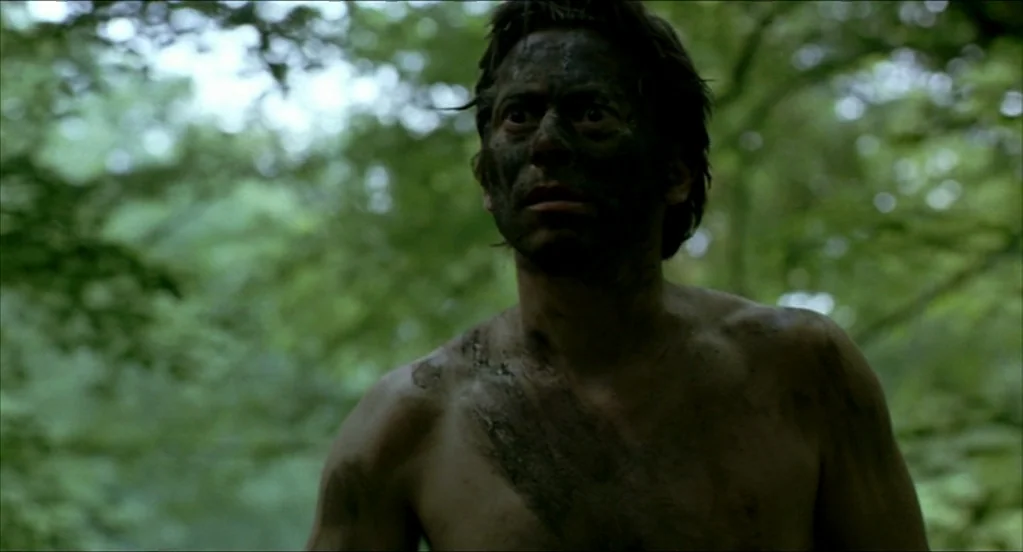 On War (2008) movie still: medium — A dense woodland with lush green canopy and blurred foliage in the background. A shirtless man with dirt-covered…