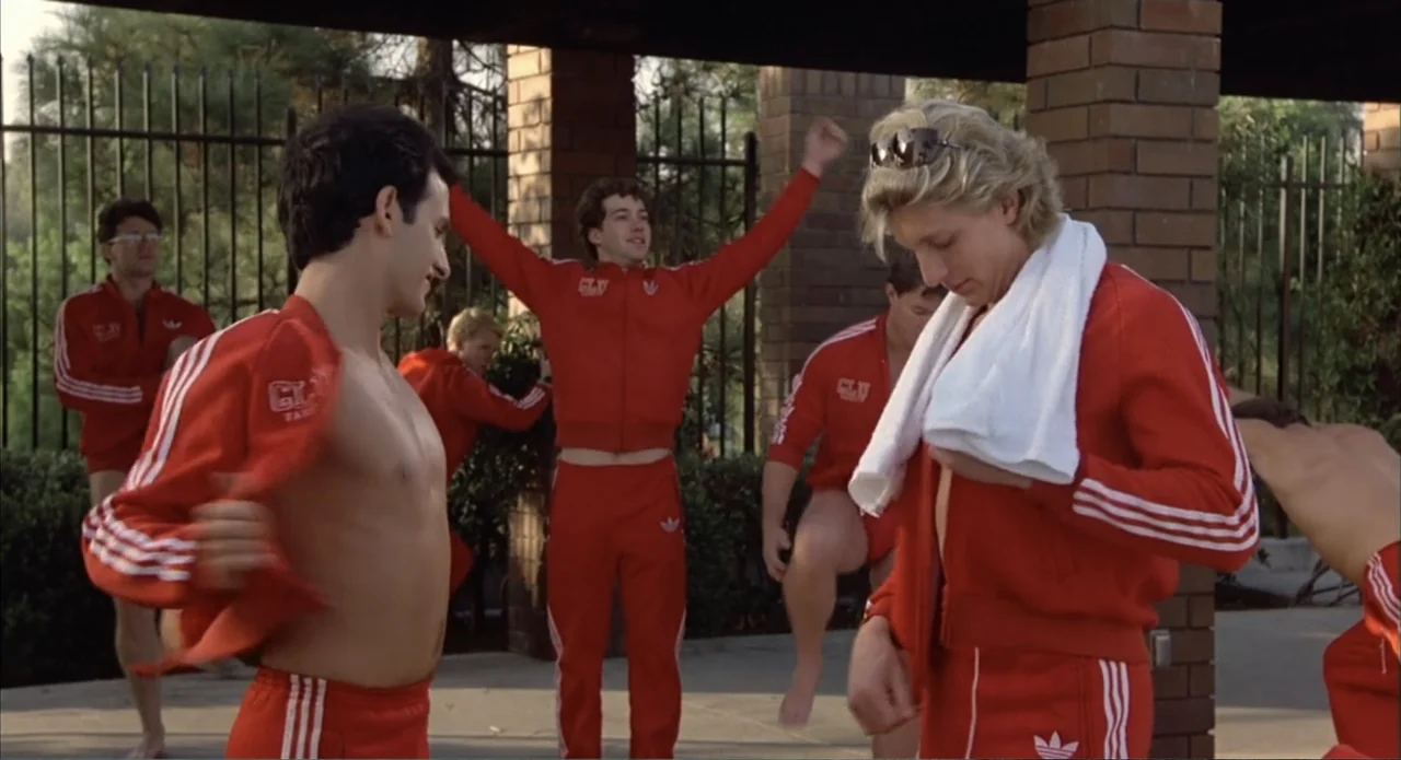 Back to School (1986) movie still: medium — A group of men stand under a brick-pillared outdoor shelter in matching red athletic wear. The men are…