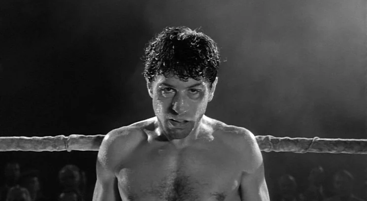 Raging Bull (1980) movie still: medium — A man stands in a dark arena inside a rope-enclosed fighting perimeter. He leans against the ropes with a…