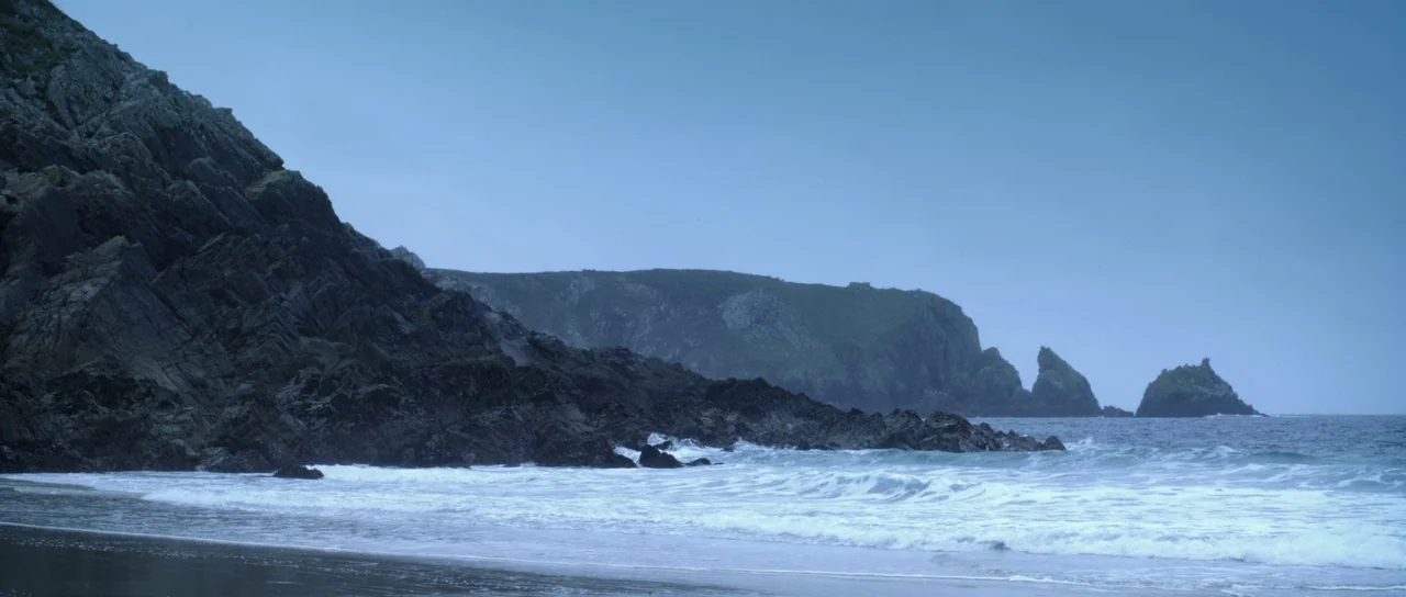 Livid (2011) movie still: establishing — A rugged rocky coastline meets the sea under a heavy, overcast blue sky. White foam rolls onto the dark…