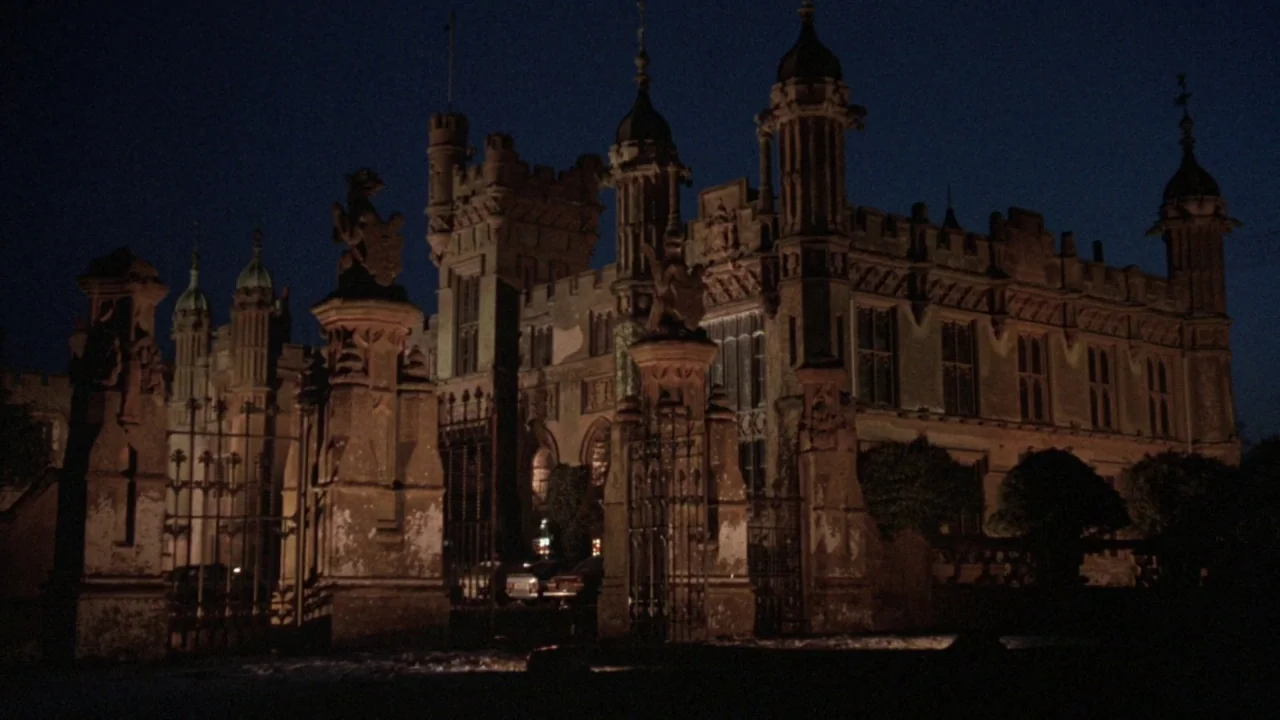 The Lair of the White Worm (1988) movie still: establishing — A dark gothic manor features pointed towers, weathered stone walls, and intricate…