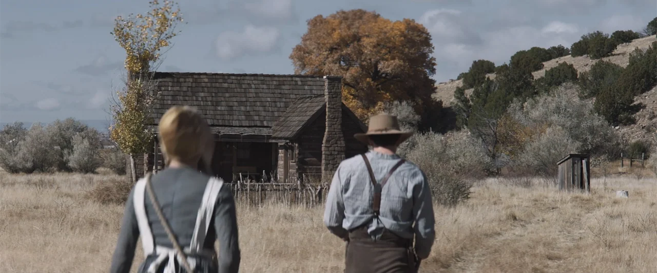 The Wind (2005) movie still: fullbody — A dry, grassy plain features a small, weathered log cabin with a stone chimney. A man and a woman walk away…