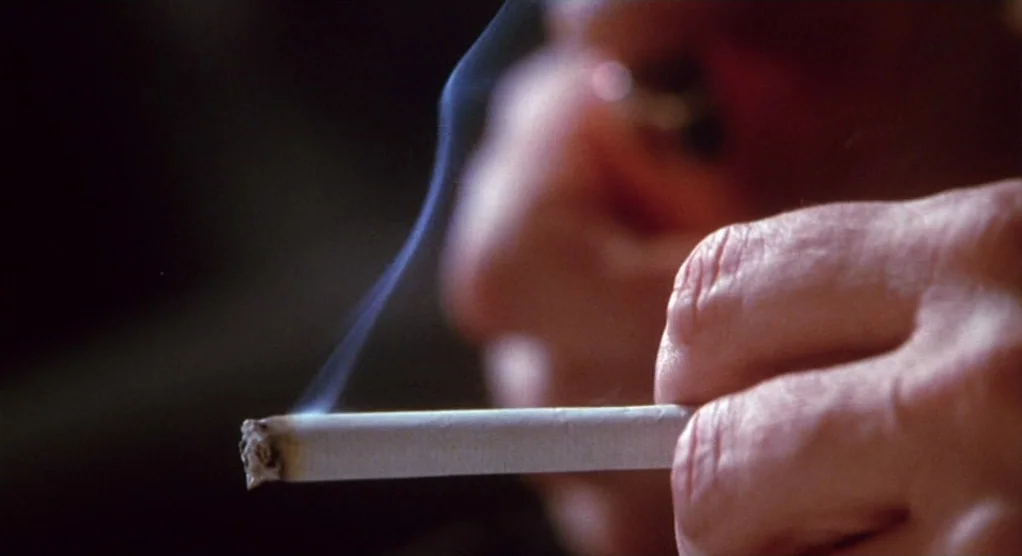 The Crossing Guard (1995) movie still: closeup — A close view shows a hand holding a lit cigarette with a thin trail of smoke rising. The subject is…