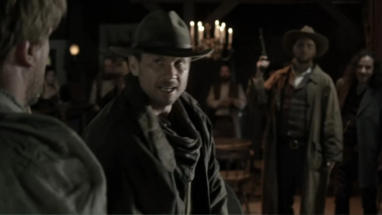 Dawn Rider (2012) movie still: medium — A dimly lit room with wood beams and a chandelier in the background. A man in a cowboy hat faces toward the…