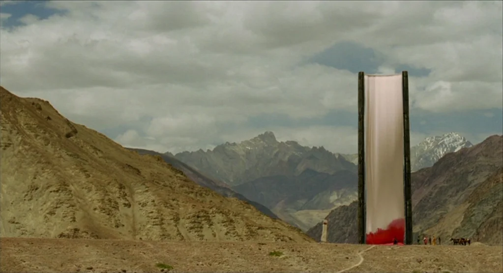 The Fall movie still: wide — A vast, arid mountain valley sits under a cloudy, overcast sky during the day. A tall, rectangular wooden frame holding…