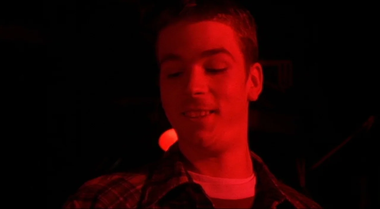 Pecker movie still: closeup — A dark space is filled with intense red light focused on a man's face. The subject is a young male wearing a plaid…