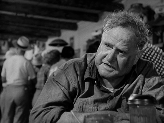 Ace In The Hole (1951) movie still: medium — A bustling interior with wooden ceiling beams and blurred figures in the background. An elderly man…