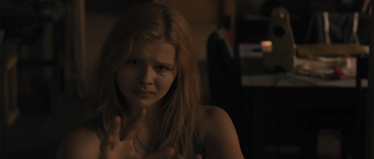 Carrie (2013) movie still: medium — A dark, dimly lit domestic space features furniture in the blurred background. A young woman with long blonde…