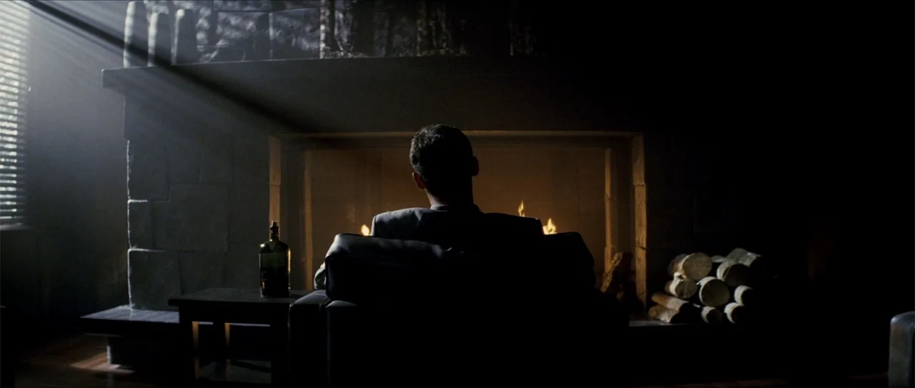 Elektra (2005) movie still: medium — A dark living room features a stone fireplace with a fire burning inside it. A man sits in a chair facing the…
