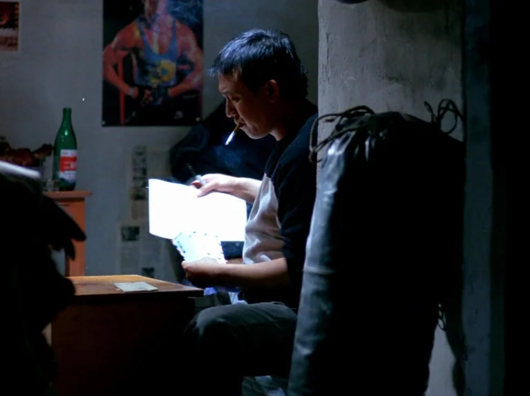 Black Snow (1990) movie still: medium — A man sits at a desk in a dark room with a poster on the wall. He holds a cigarette in his mouth while…