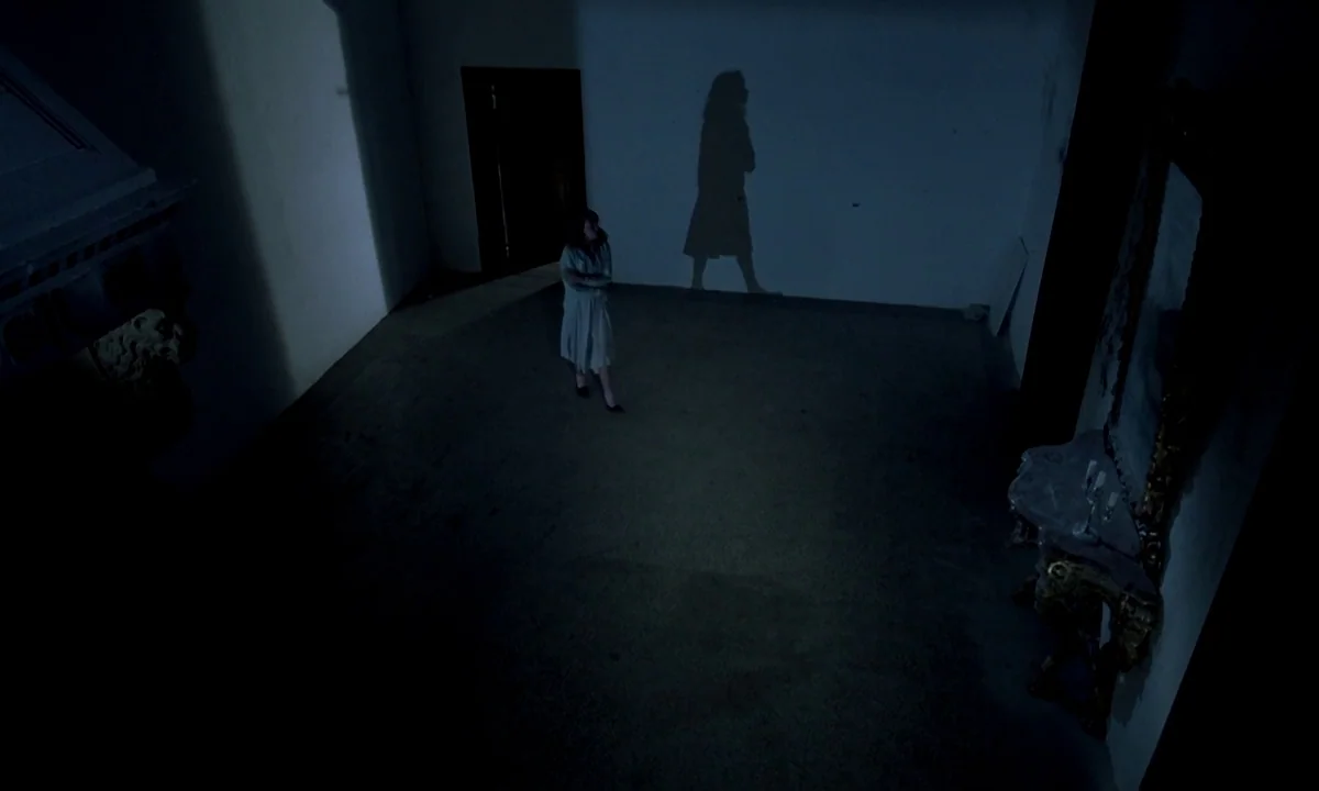 Phenomena (1985) movie still: wide — A large, sparsely furnished hall with smooth walls and dark corners. A woman stands alone while her large…
