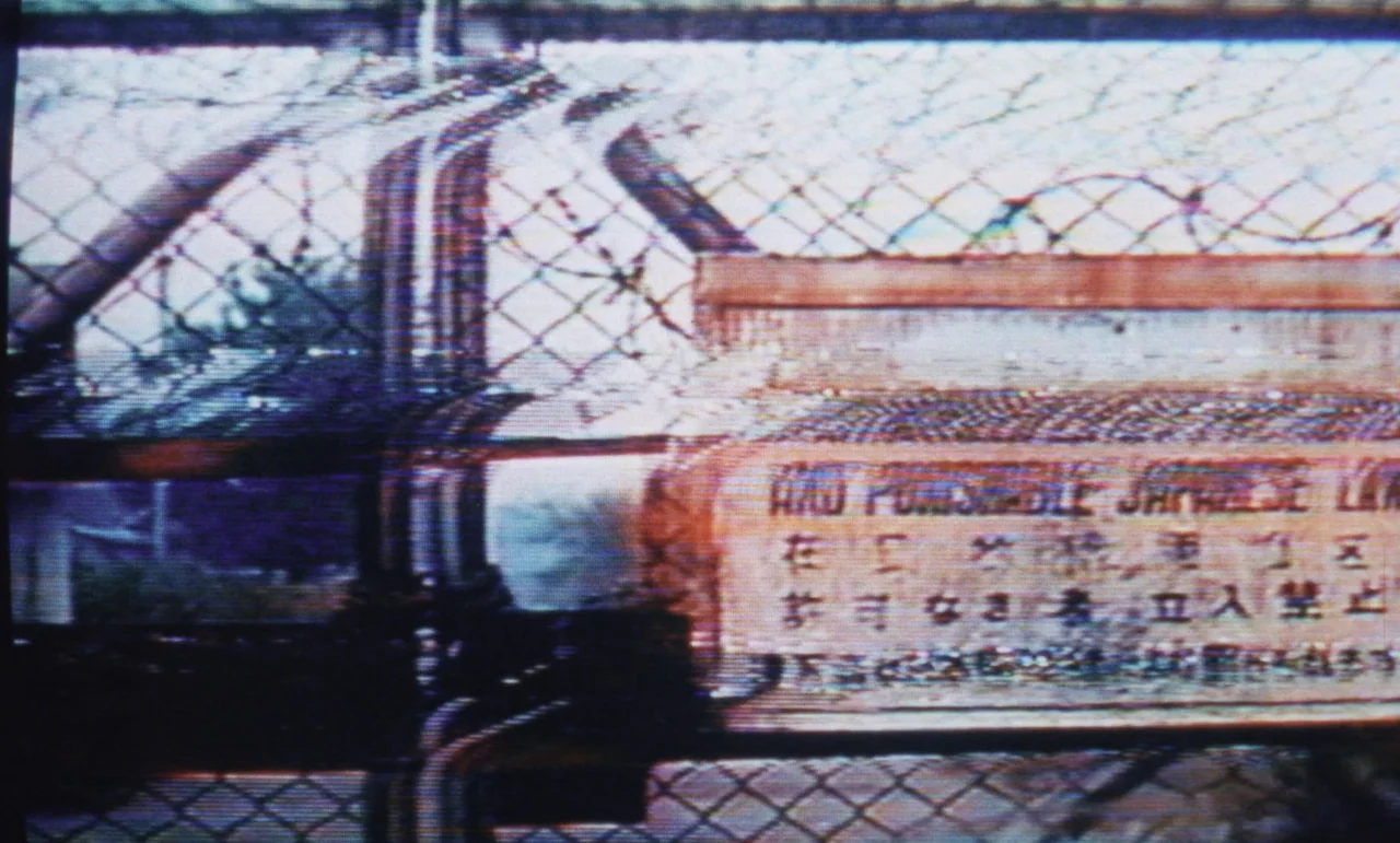 Evil Dead Trap (1988) movie still: closeup — A metal chain-link fence has a rectangular sign with English and Japanese text attached. The sign warns…