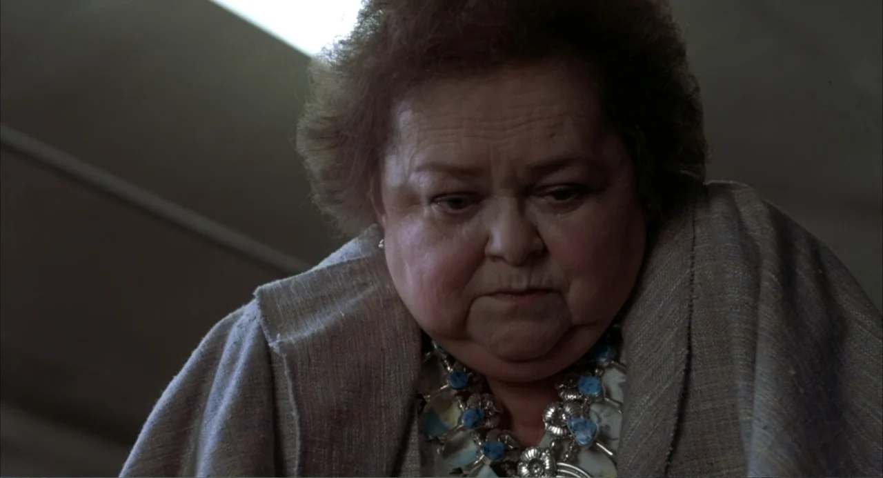 Poltergeist III (1988) movie still: closeup — An interior space with neutral walls and soft top-down lighting. An elderly woman looks downward…