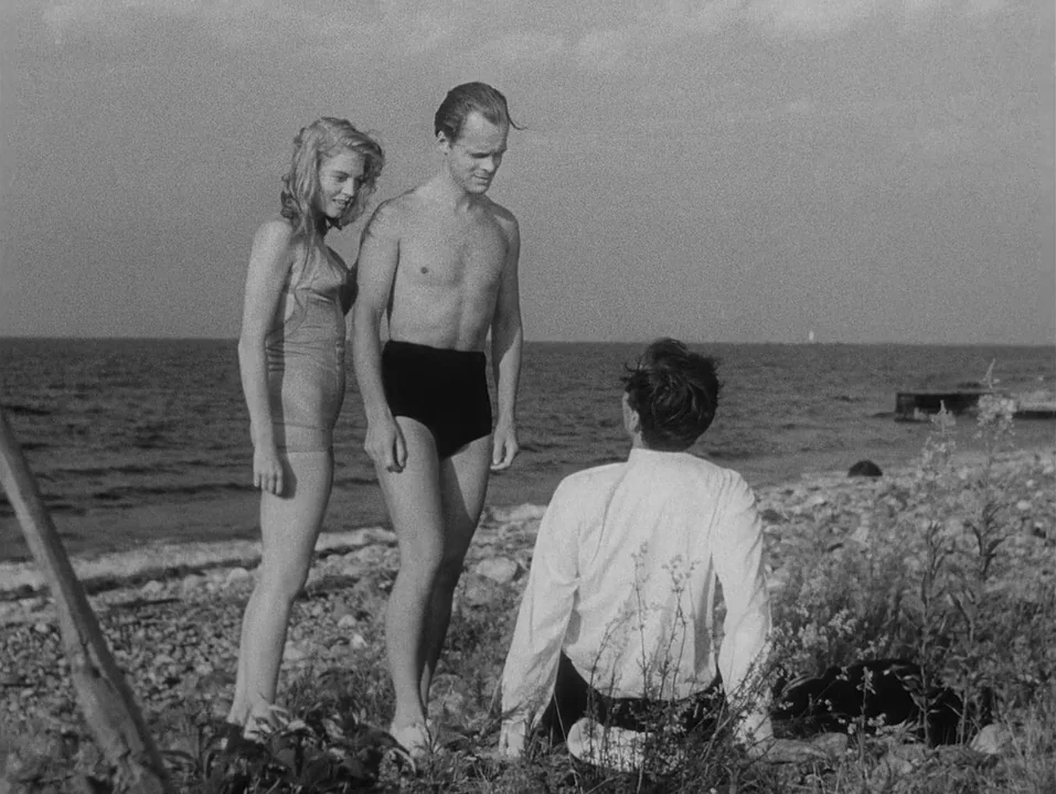A Ship Bound for India (1947) movie still: fullbody — A pebbled beach with sparse vegetation borders a calm sea under an overcast sky. A woman and a…