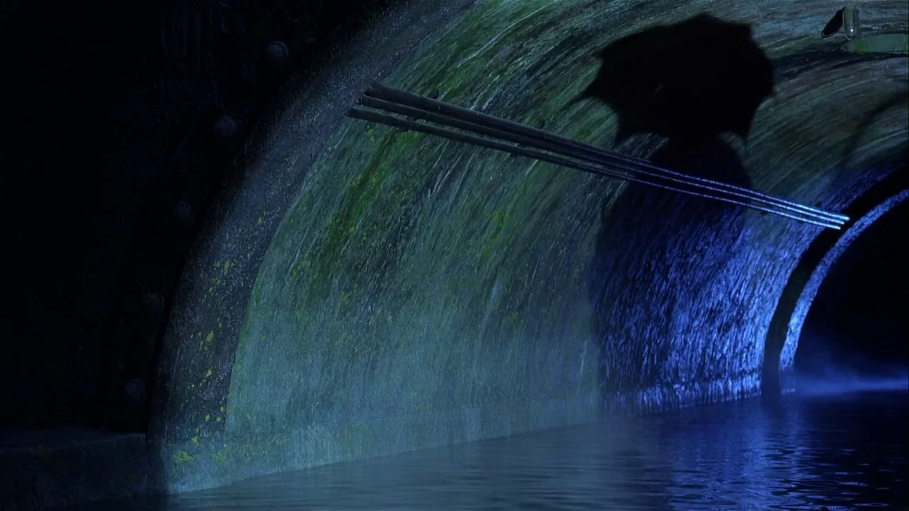 Batman Returns (1992) movie still: wide — A dark subterranean tunnel features a curved brick wall covered in patchy green moss. The elongated shadow…