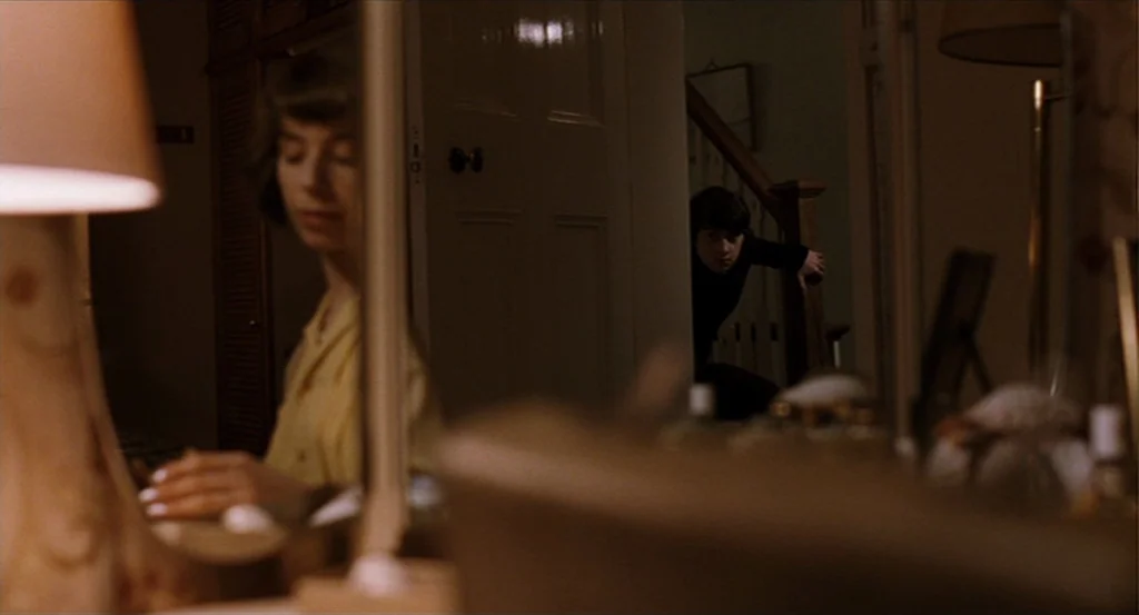 Submarine movie still: medium — A dimly lit room features a large mirror reflecting a woman and a doorway. A young man stands in the dark hallway…