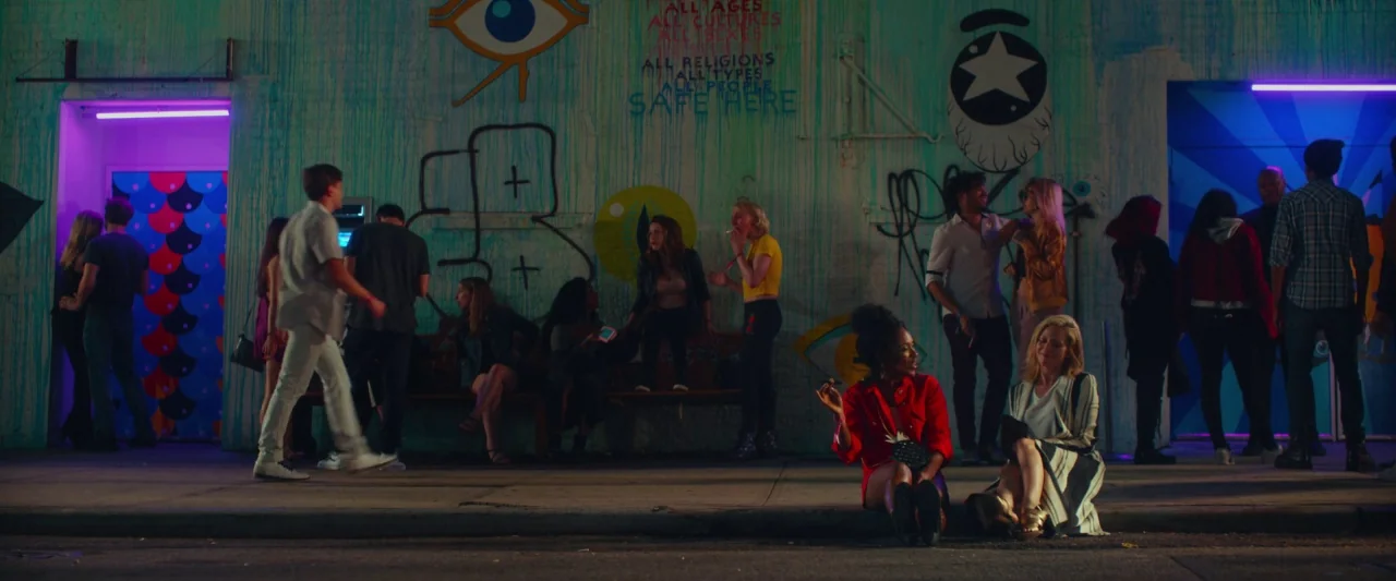 Someone Great (2019) movie still: wide — A graffiti-covered wall serves as a backdrop for a group of people at night. The wall features text and art…