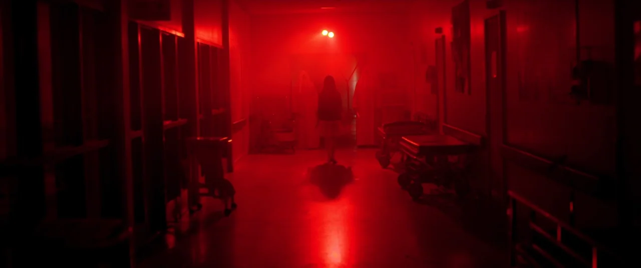 The Void (2016) movie still: wide — A long, dimly lit hospital corridor bathed in intense red light. A young girl walks away from the camera toward…