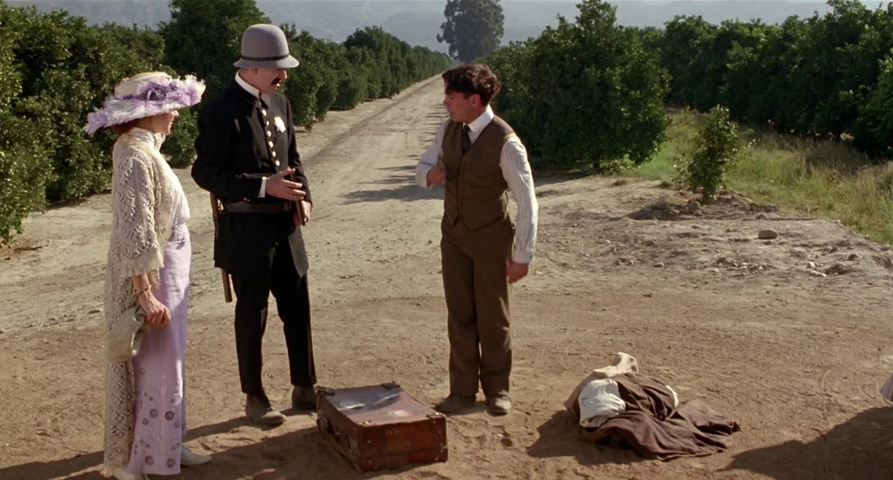 Chaplin (1992) movie still: fullbody — A dirt path cuts through an expansive orchard with rows of green fruit trees. Three people in mid-century…