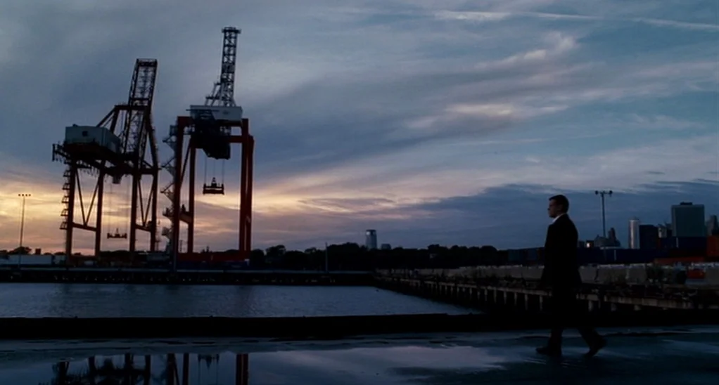 The Adjustment Bureau (2013) movie still: wide — A vast industrial shipping dock features two large orange cranes against a dusk sky. A man walks in…