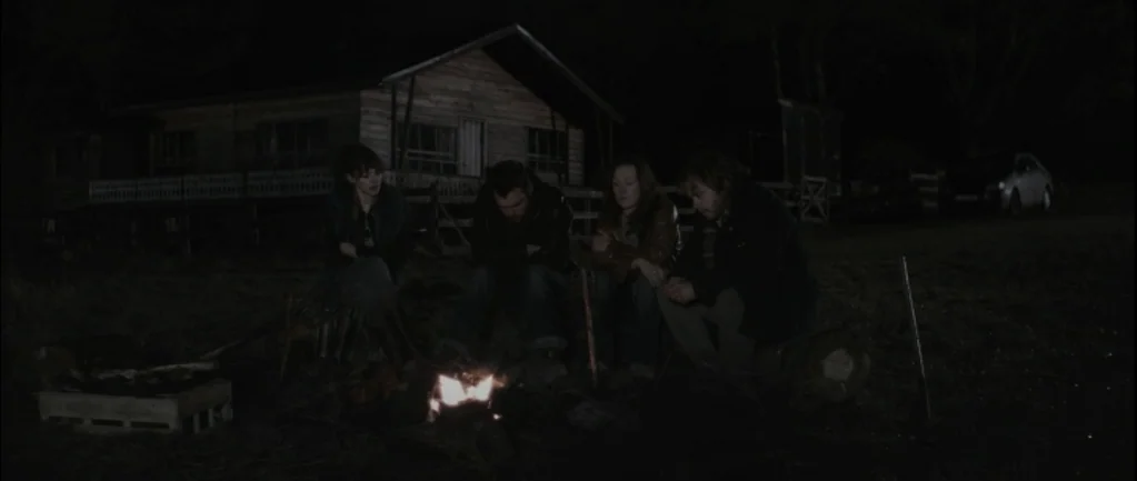 One Hundred Mornings (2009) movie still: wide — Four people sit on logs in front of a small fire in a dark field. Behind them stands a wooden house…