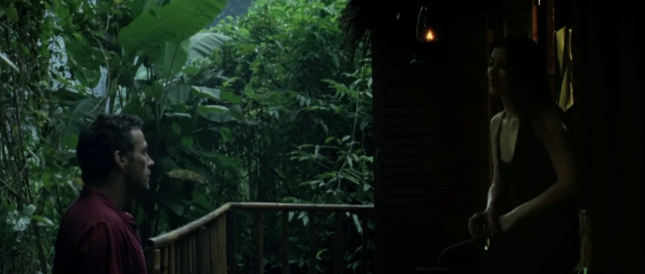 Vinyan (2008) movie still: wide — A wooden balcony overlooks dense tropical foliage during the evening. A man in a red shirt faces a woman sitting…