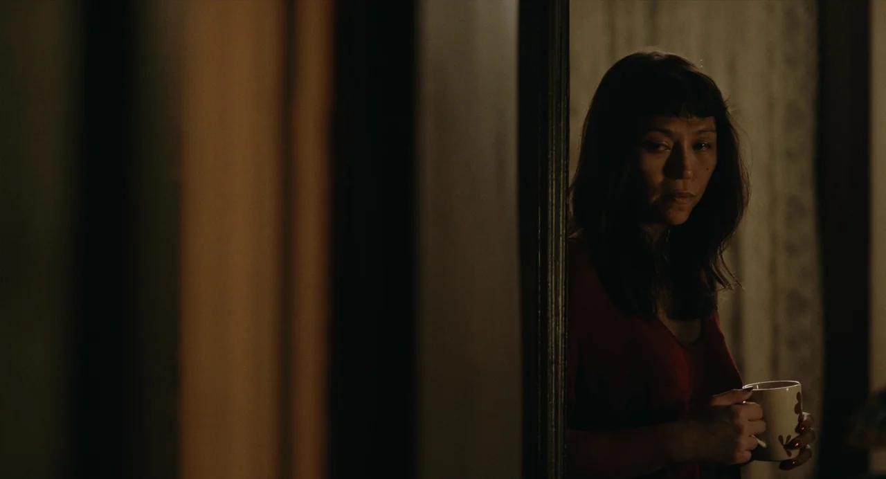 Lingua Franca (2019) movie still: medium — A woman stands in a dimly lit doorway with a wooden frame visible. She holds a patterned mug in both…