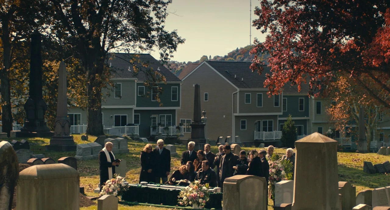 A Beautiful Day In The Neighborhood movie still: wide — A cemetery with scattered stone markers is situated in front of modern suburban houses. A…