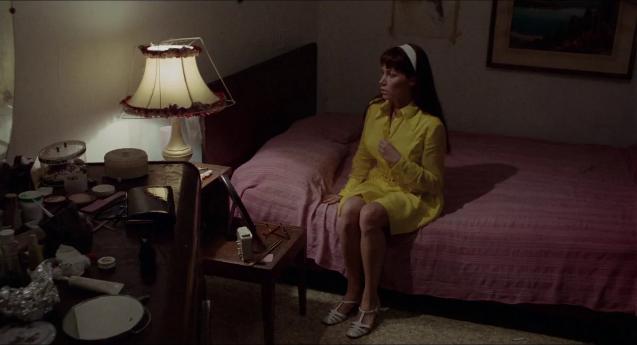 Medium Cool (1969) movie still: medium — A dimly lit room with a single bed and a cluttered dressing table. A woman sits on the edge of the mattress…