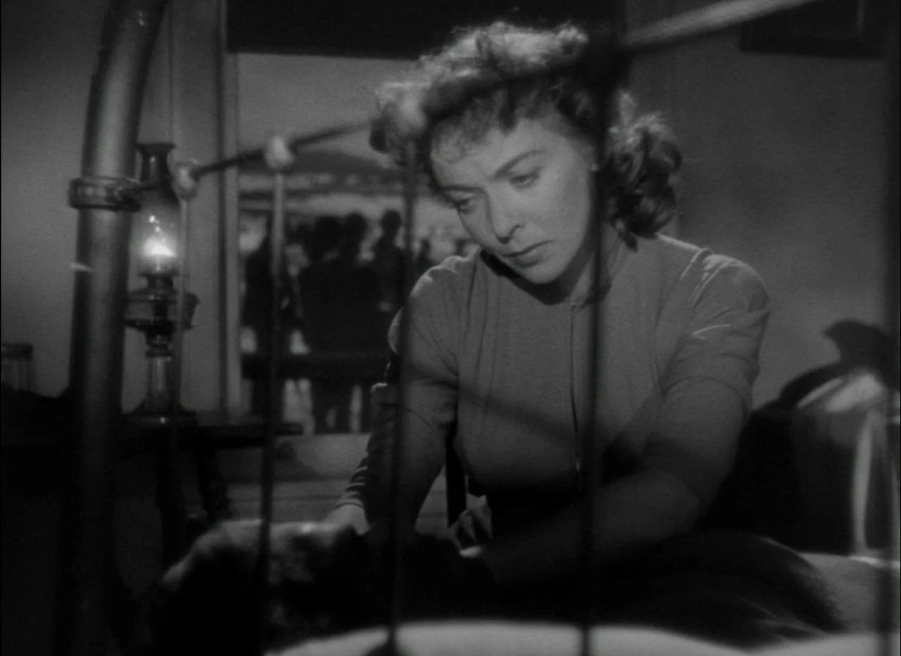 On Dangerous Ground (1951) movie still: medium — A dimly lit room contains an oil lamp and an iron-framed bedstead. A woman leans over a person…