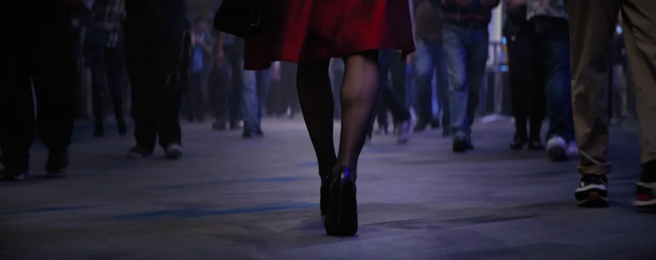 The Naked Gun (2025) movie still: closeup — A low-angle view shows the legs of a woman walking through a busy space. The focus remains on her red…