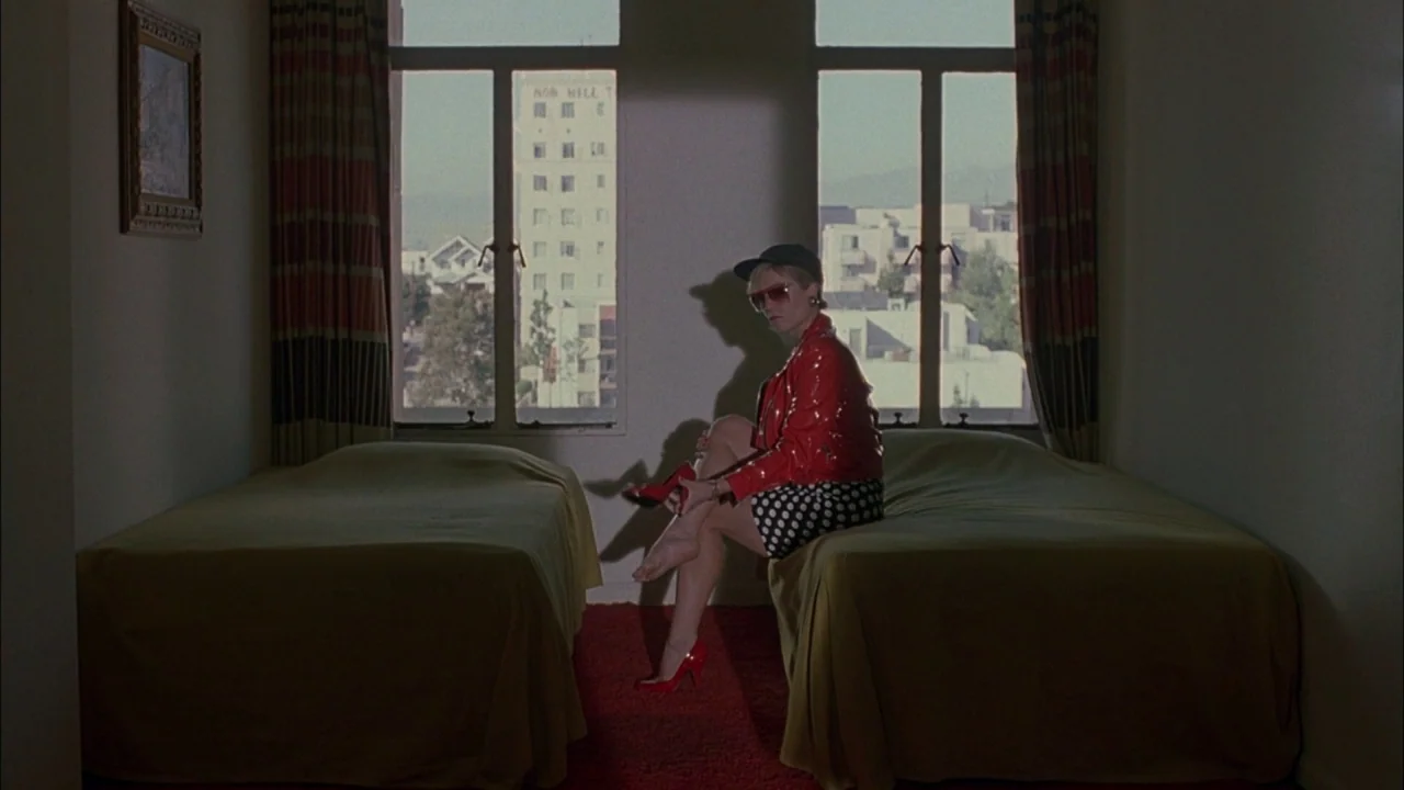 Whore (1991) movie still: wide — A spare motel room with two twin beds features large windows overlooking a city street. A woman sits on the edge of…