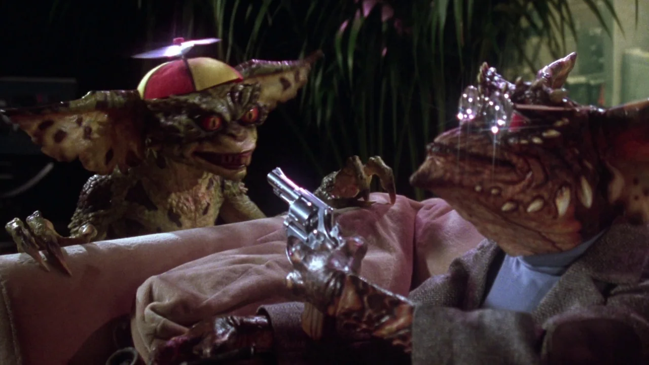 Gremlins 2: The New Batch movie still: medium — Two reptilian creatures sit on a cushioned surface in a dimly lit space. One creature points a small…