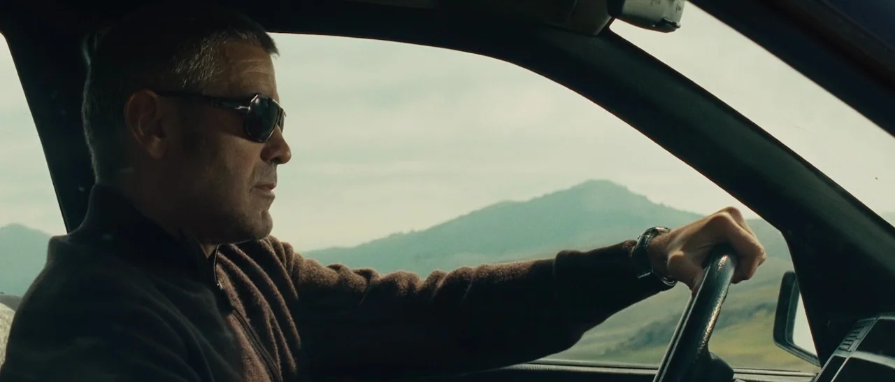 The American (2010) movie still: medium — The interior of a car driving through a hilly landscape under an overcast sky. A man wears sunglasses and…