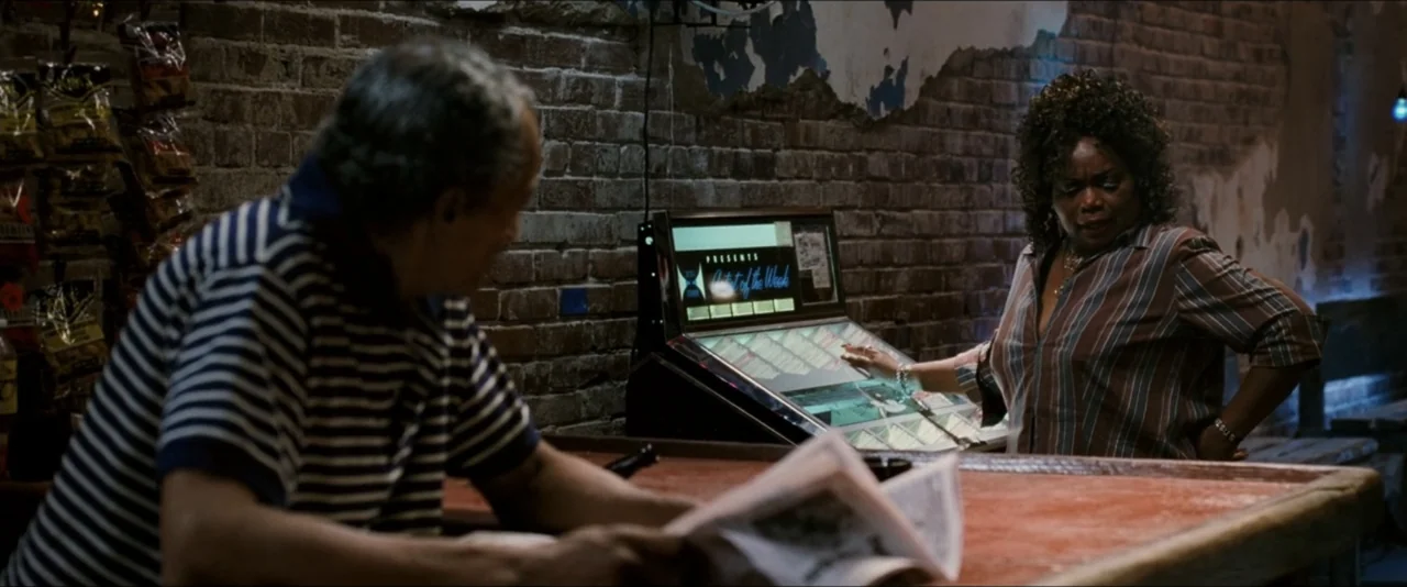 Black Snake Moan (2006) movie still: medium — A dimly lit room features a worn brick wall with patches of white plaster. A man sits at a table…