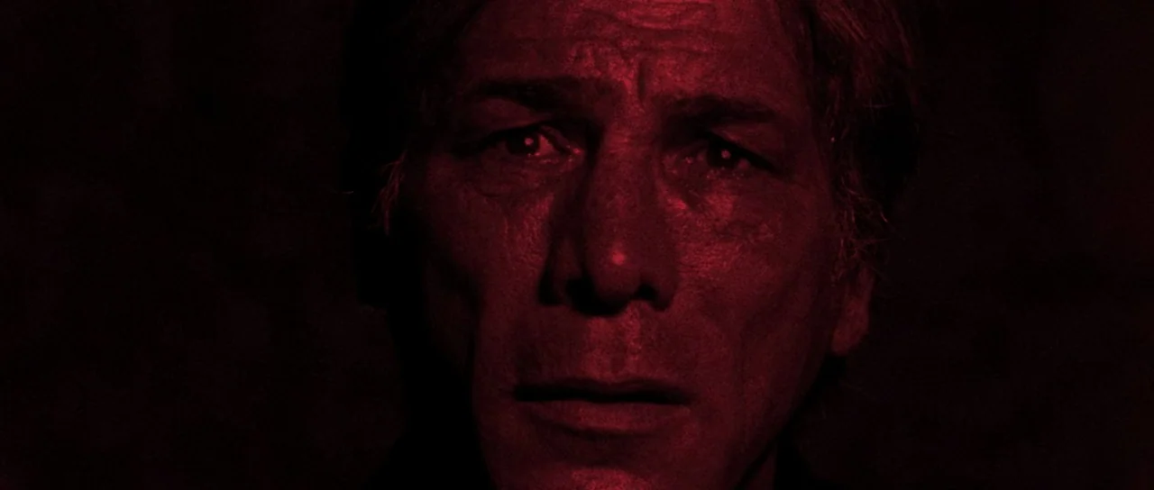 Let The Corpses Tan (2017) movie still: closeup — A man is positioned in a dark, shadow-filled environment with no background detail visible. The…