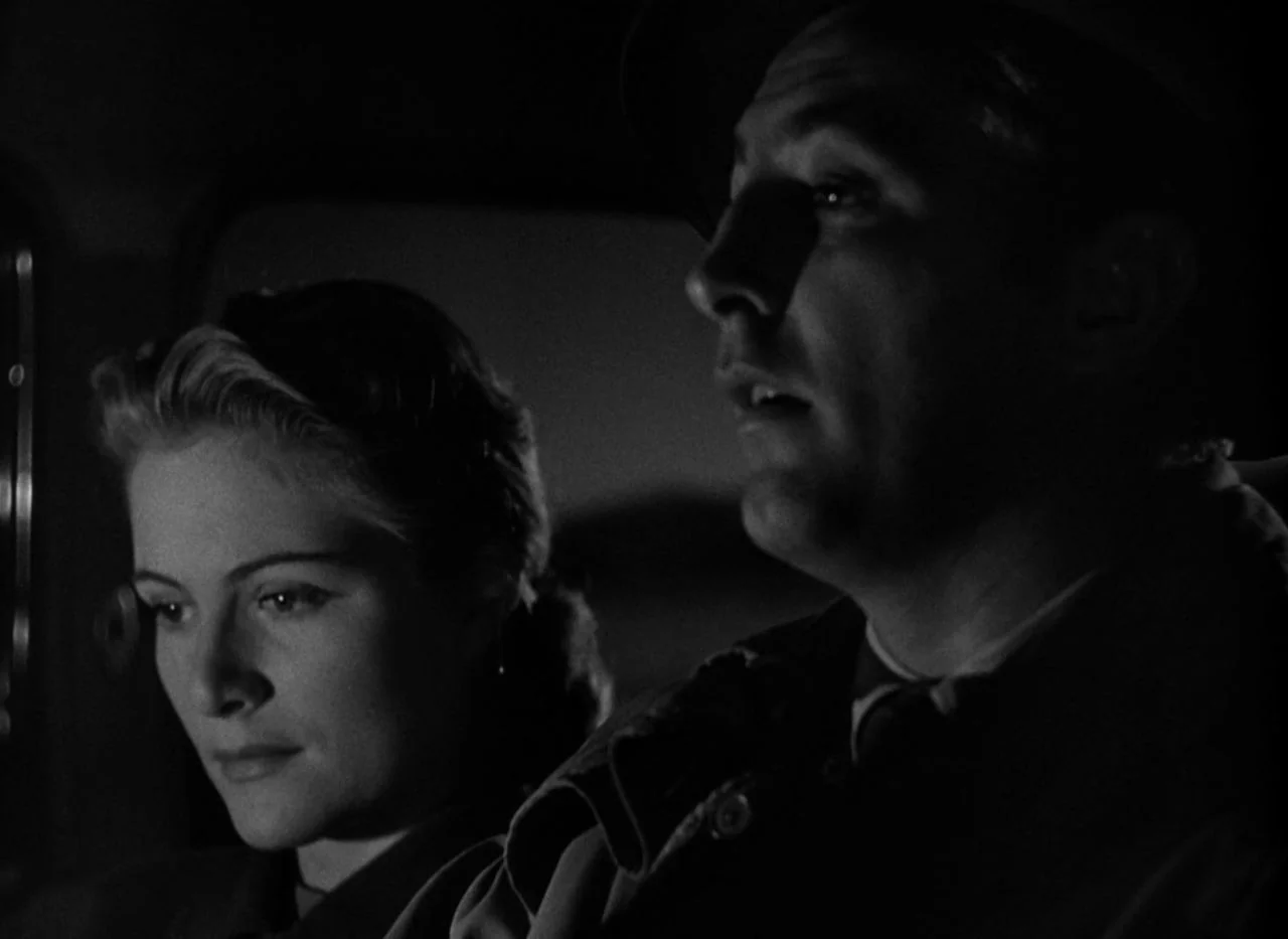 Out of the Past (1947) movie still: medium — The interior of a moving car at night is dimly lit with deep shadows. A man operates the steering wheel…