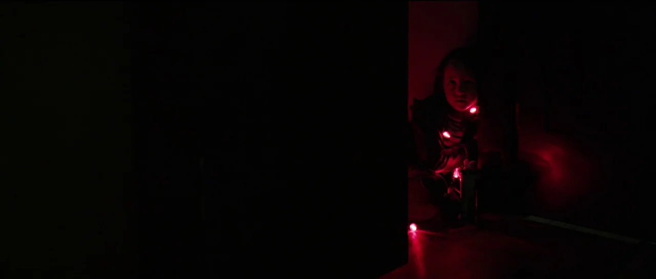 The Boogeyman (2023) movie still: medium — A dark space defined by large, obscured shapes of furniture or doors. A small child sits in the narrow…