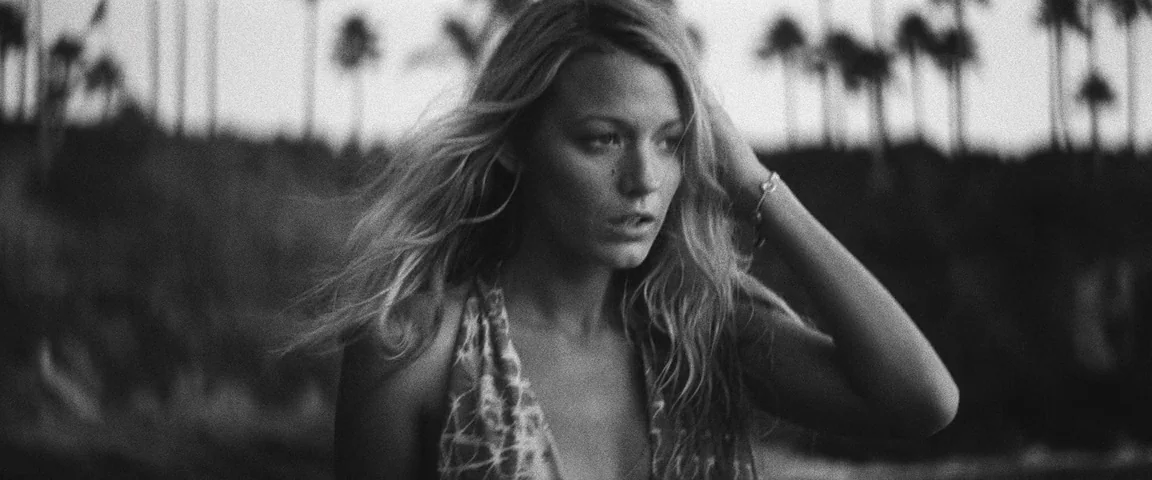 Savages (2012) movie still: medium — A woman stands in front of a dark, out-of-focus background of palm trees. She is adjusting her hair with one…