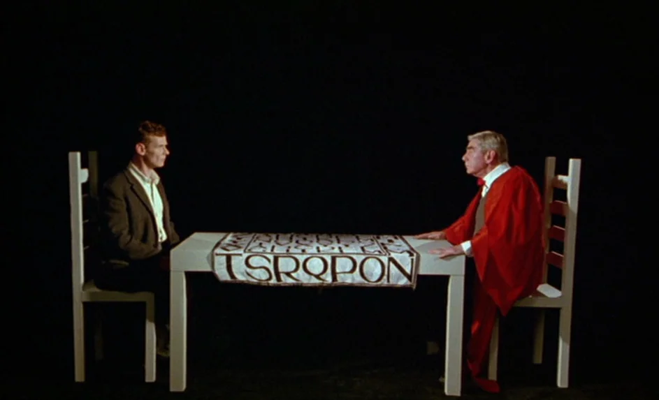 Wittgenstein (1993) movie still: wide — Two men sit at a plain white table against a completely black background. A white cloth with text drapes…