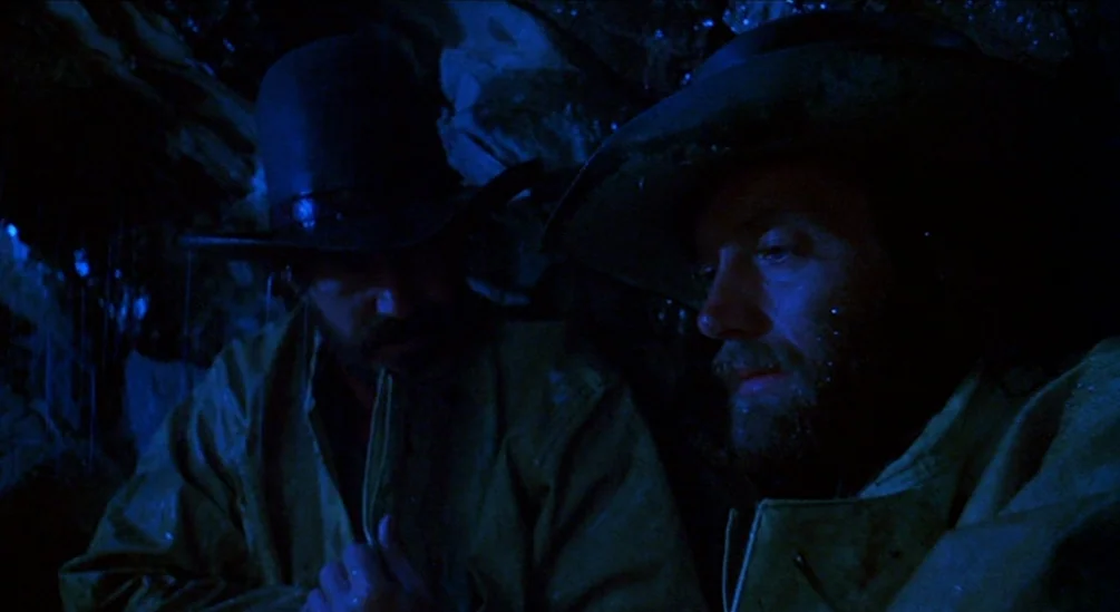 The Hired Hand (1971) movie still: medium — Two men sit under cover in a wet, dark outdoor environment during a downpour. Both men wear hats and…